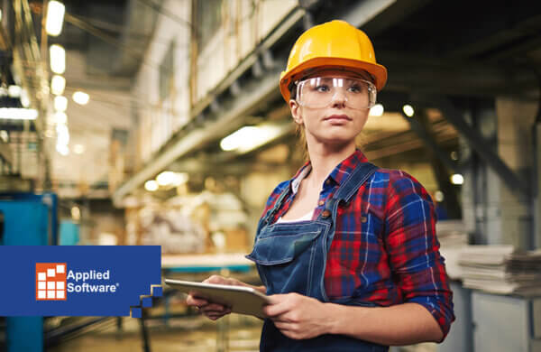 在工厂模糊的背景下，一名女子手持移动设备，戴着黄色安全帽和安全眼镜