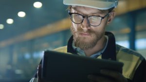 男人的胡子戴着黑边的眼镜,白色安全帽看着电子平板电脑,背景模糊的工厂有四个白色的天花板灯