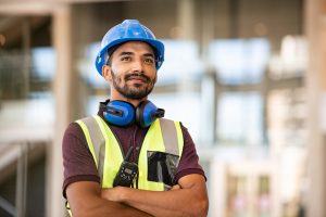 大胡子的照片建筑工作工人穿栗色马球衬衫,黄色的安全背心和蓝色安全帽,蓝色的听力保护耳机在脖子上,模糊背景的建筑工地